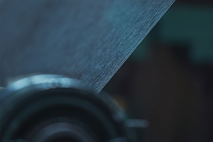 This close-up abstract image features a textured, dark blue or grey surface angled across the frame, likely a fabric or industrial material. It appears to be moving over a blurred, dark circular object, suggesting a process of winding or feeding material