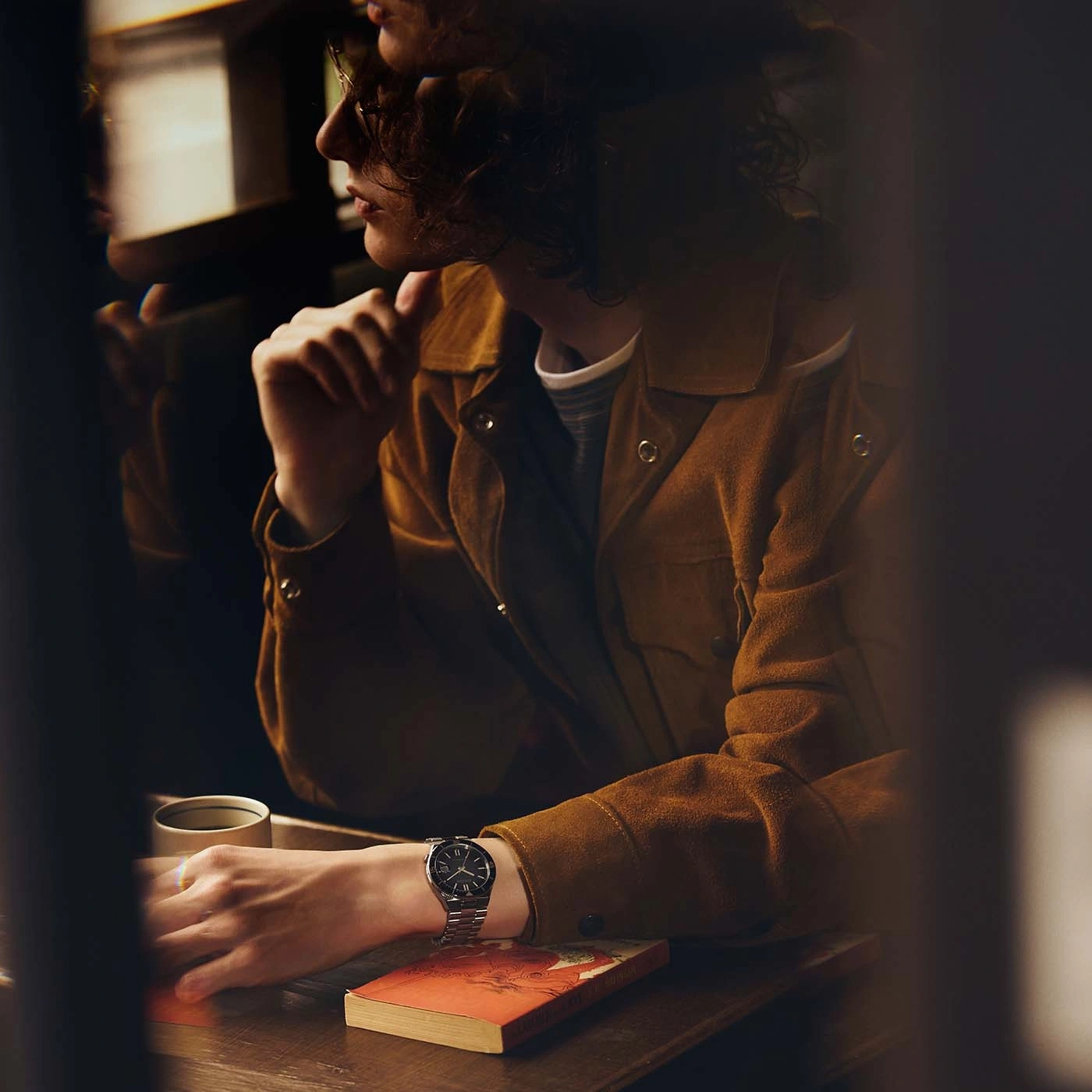 Nahaufnahme einer Person mit Uhr und Jacke, die an einem Tisch mit einem Buch sitzt.