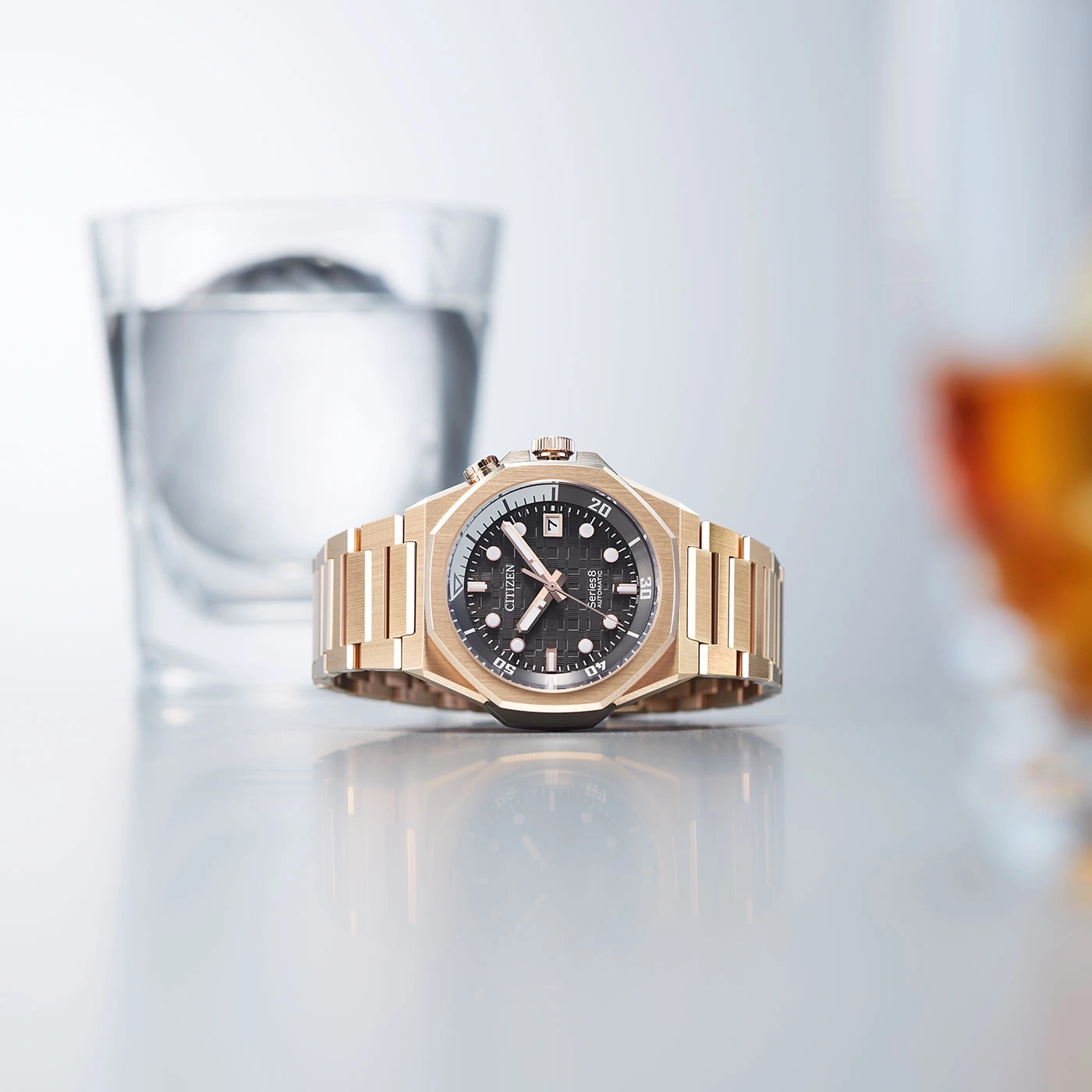 Goldene Uhr mit schwarzem Zifferblatt, Glas Wasser und Reflexion auf der Oberfläche.
